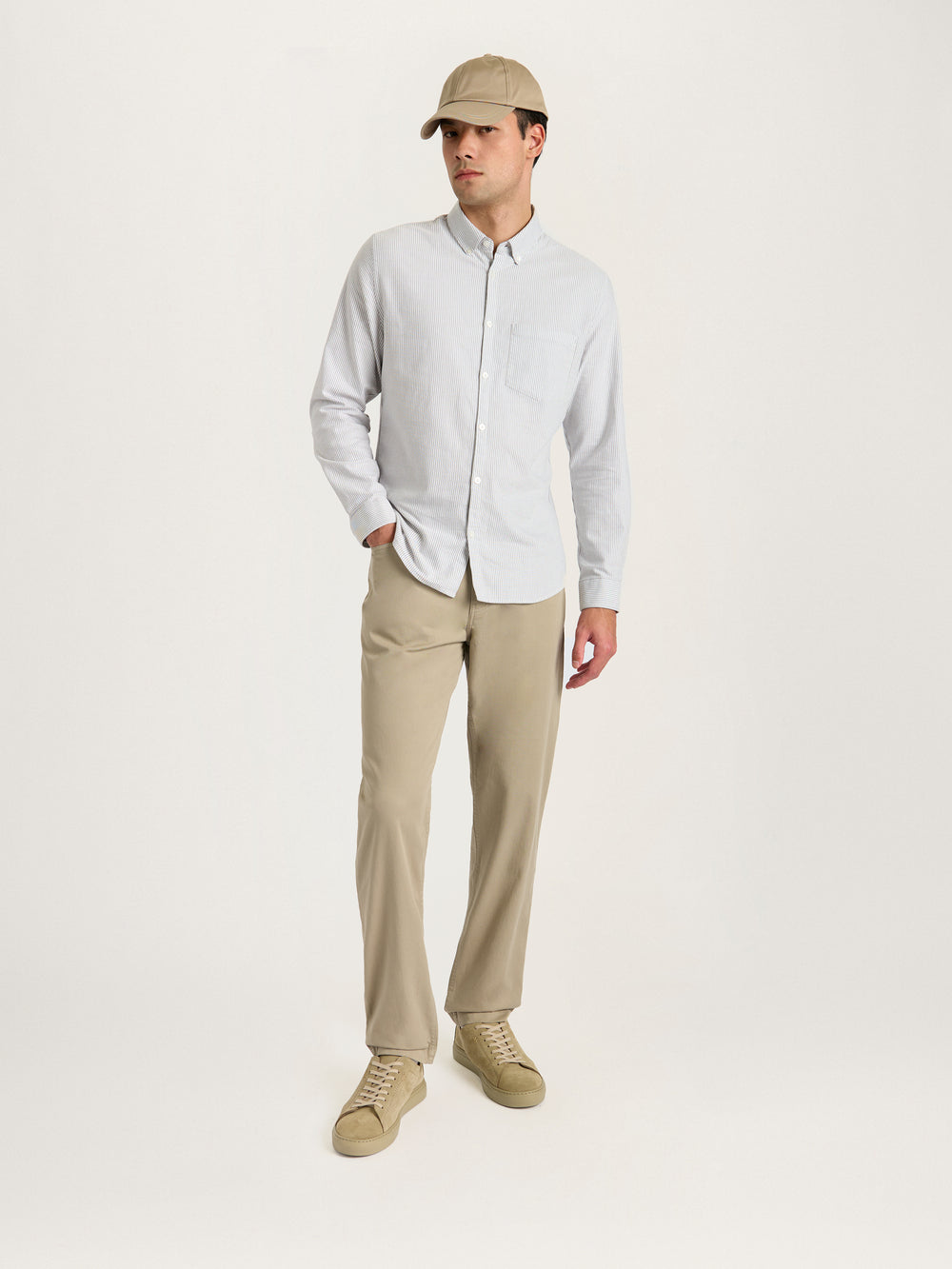 Model wearing Lestrange 5-pocket trouser in beige paired with striped button-down shirt, beige cap, and sneakers, full-length lifestyle shot.