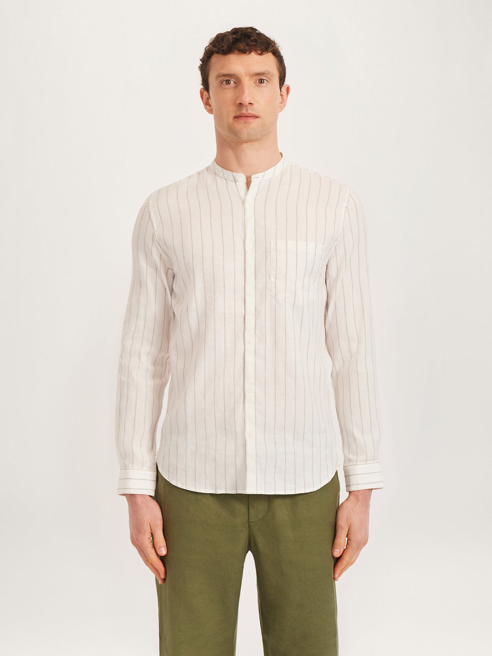 A man wearing a beige collarless linen shirt with ecru stripes and green trousers.