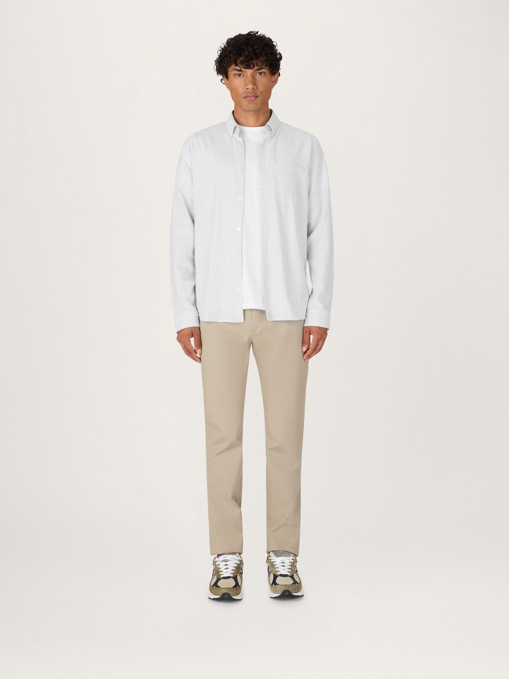 A person wearing a classic grey striped Oxford shirt with collar, buttoned up to the neck, paired with beige trousers and standing against a white background.