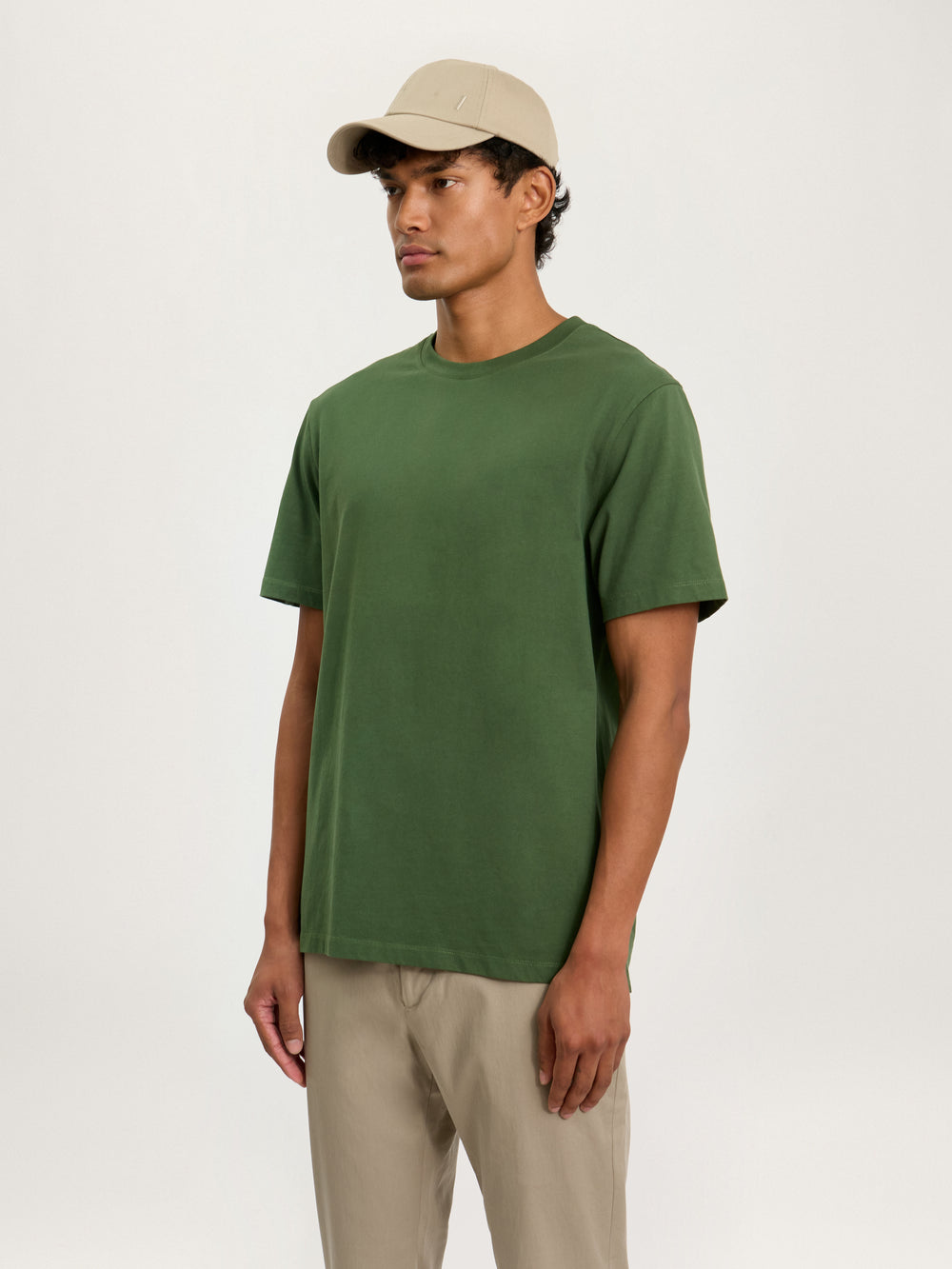 A person wearing a green heavyweight organic cotton t-shirt with ribbed neck, paired with beige pants and a beige cap.