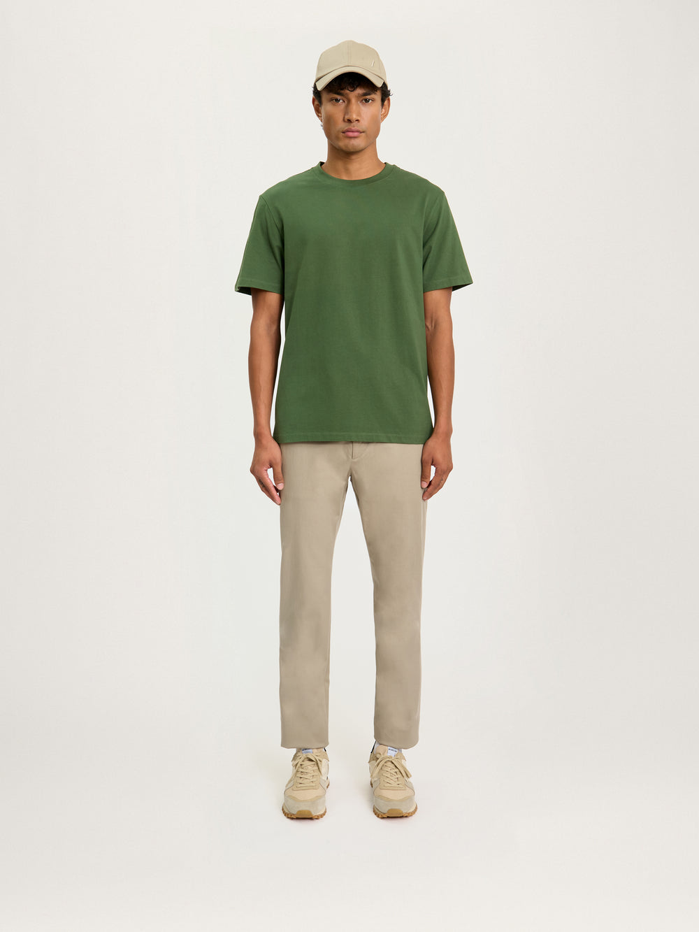 A person standing against a white background, wearing a relaxed fit olive green t-shirt and light beige trousers, paired with white sneakers.