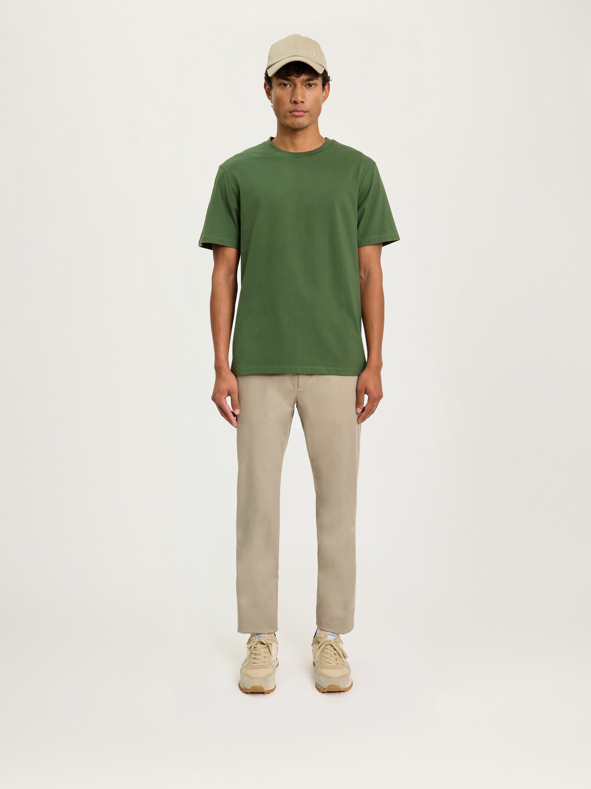 A person standing against a white background, wearing a relaxed fit olive green t-shirt and light beige trousers, paired with white sneakers.
