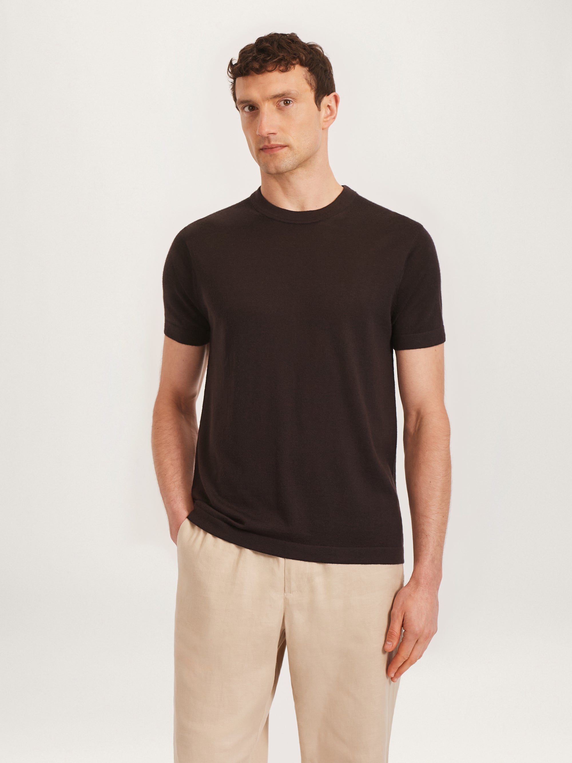 A man wearing a short-sleeved black merino wool T-shirt and beige pants standing against a white background.