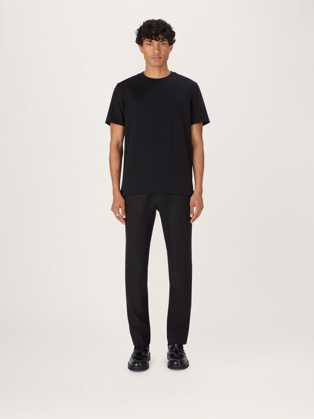 A person standing against a neutral background, wearing a black, short-sleeved, crew-neck t-shirt and dark trousers.