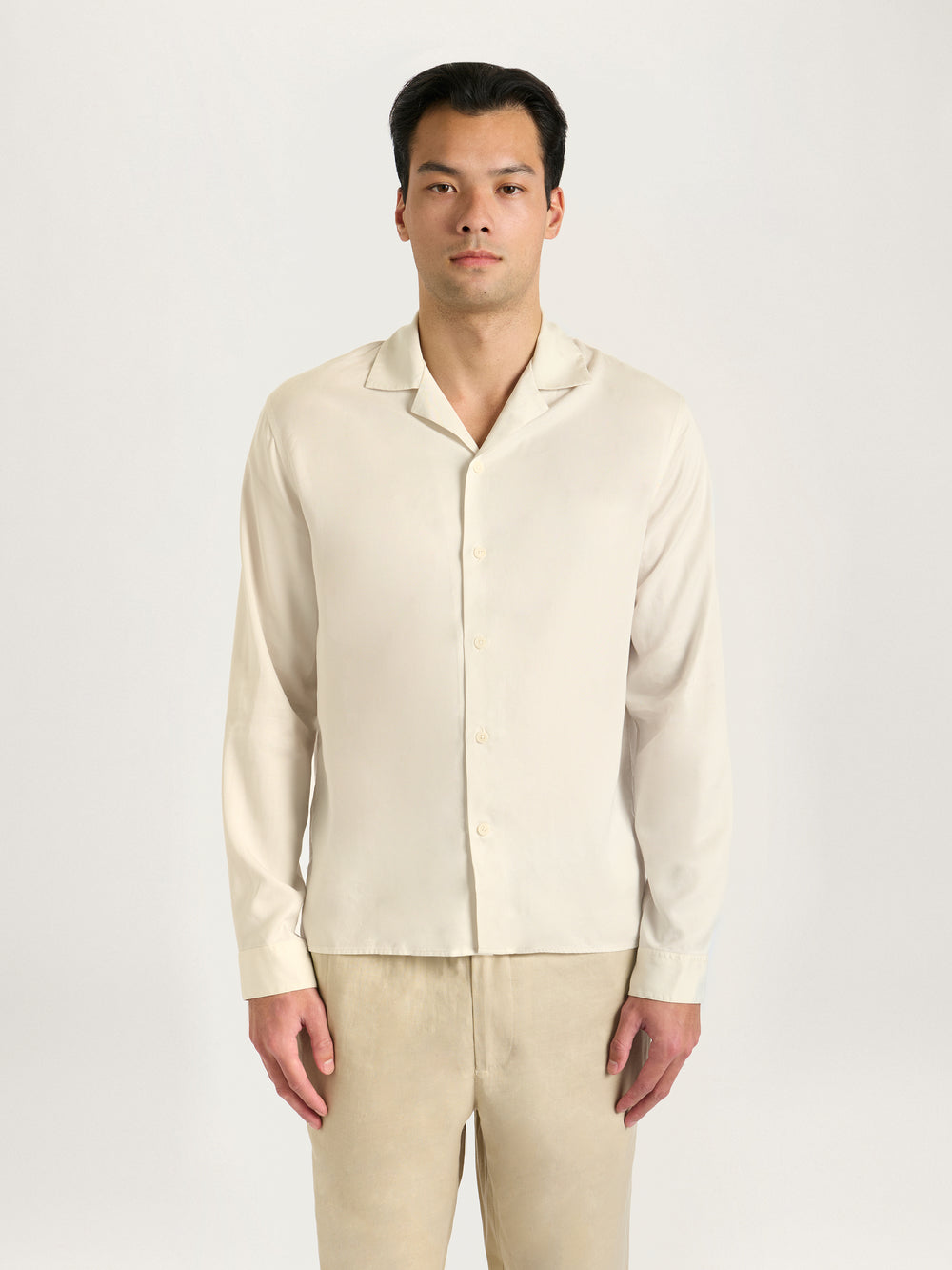 A male model wearing a beige long sleeve shirt with a Cuban collar and matching beige trousers.