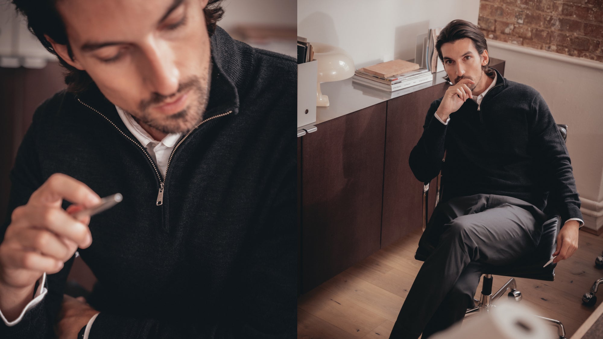 Man in smart casual outfit sitting in modern office, wearing a black quarter-zip sweater over a white shirt and grey trousers, thoughtful and holding a pen