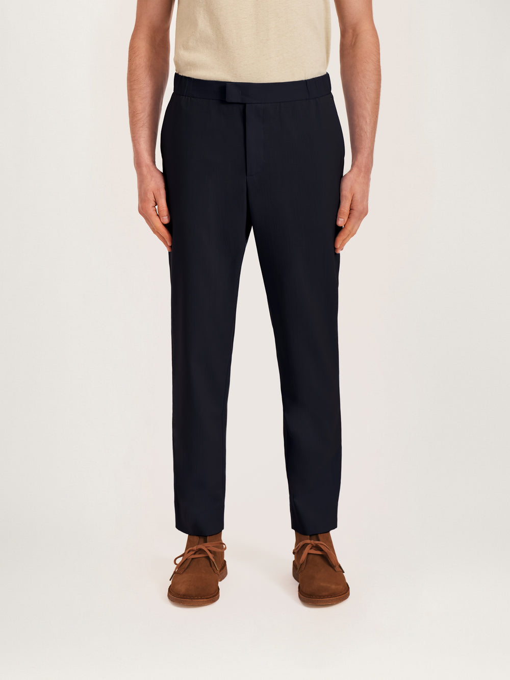 A male model wearing navy blue tropical wool trousers with a relaxed fit and a hidden elastic waistband, paired with a light-colored shirt and brown shoes.