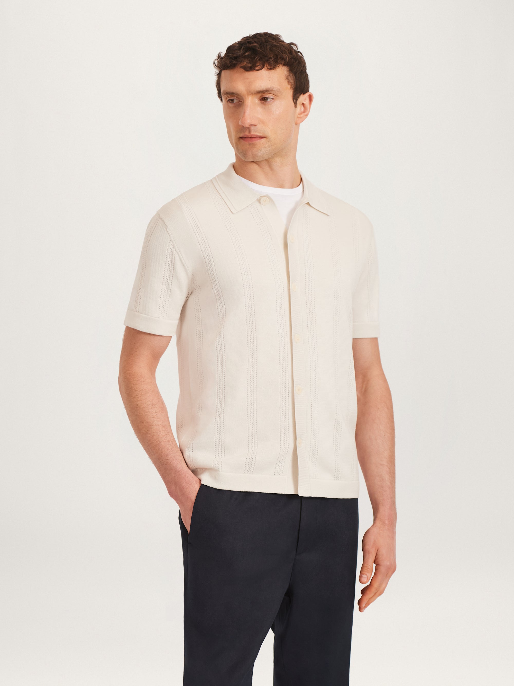 A man wearing a short-sleeve buttoned knit shirt in ecru color, with the shirt collar visible and the sleeves buttoned.