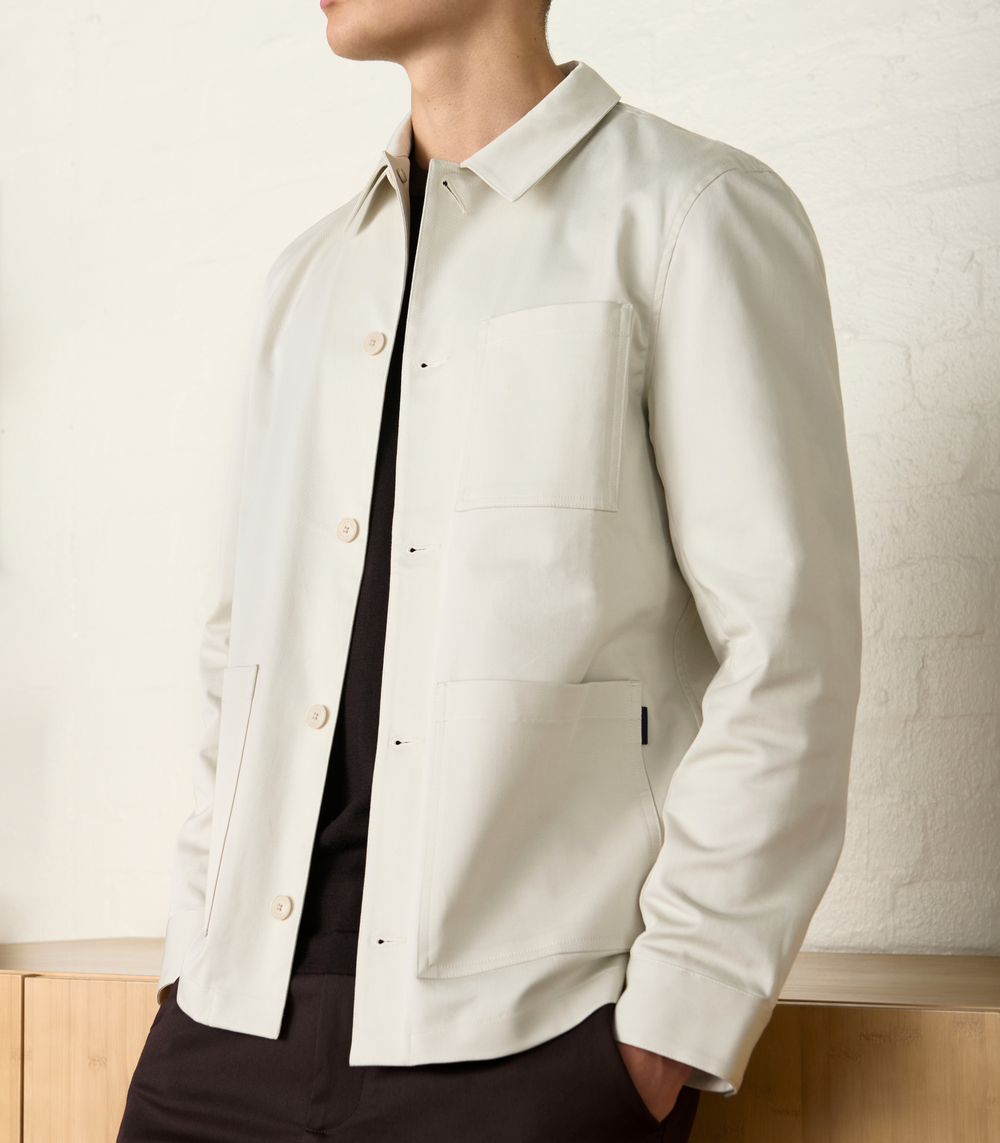 Men’s minimalist off-white button-up jacket with chest and front pockets, styled with a black shirt and dark trousers – modern casual outfit.