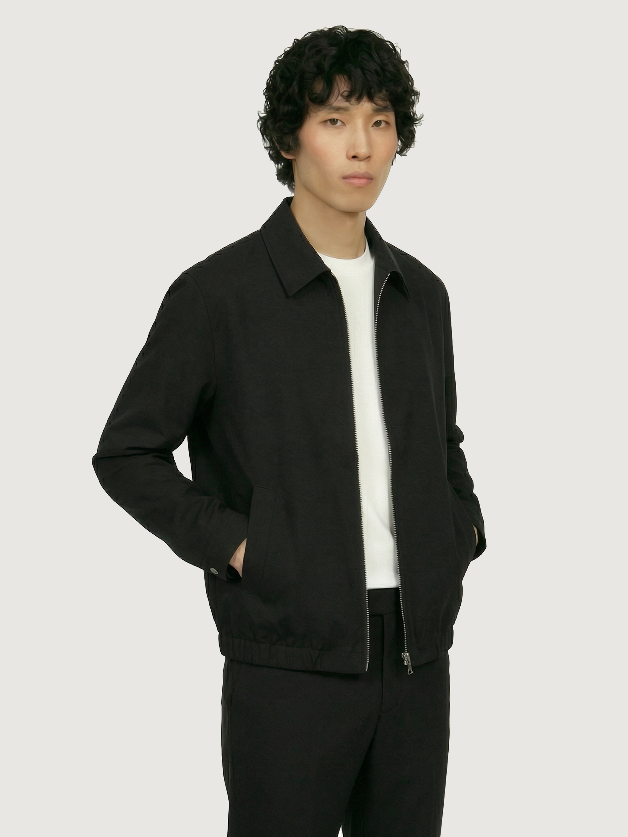 Male model wearing The 24 Coach Jacket in black tropical wool with matching black trousers and a white T-shirt.