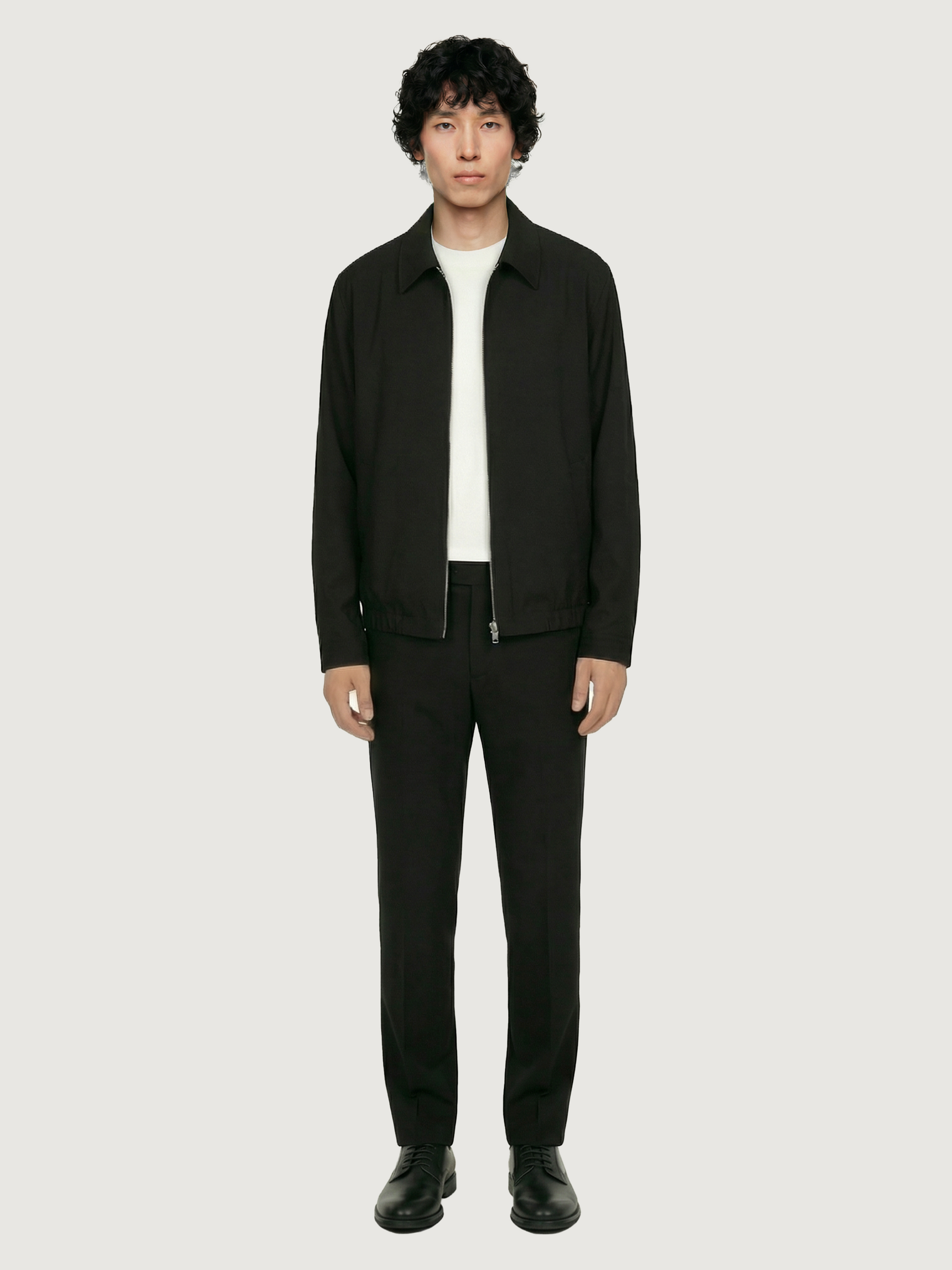 Front view of The 24 Coach Jacket in black tropical wool, styled with matching black trousers, black shoes and a white T-shirt.