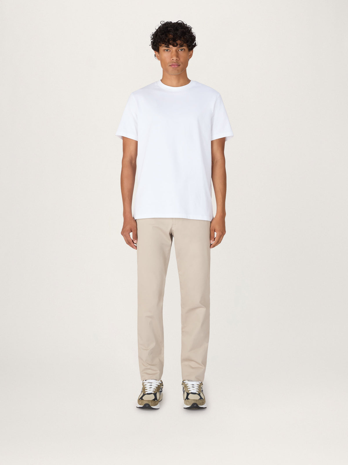 A person standing against a neutral background, wearing a white classic tee and beige pants.