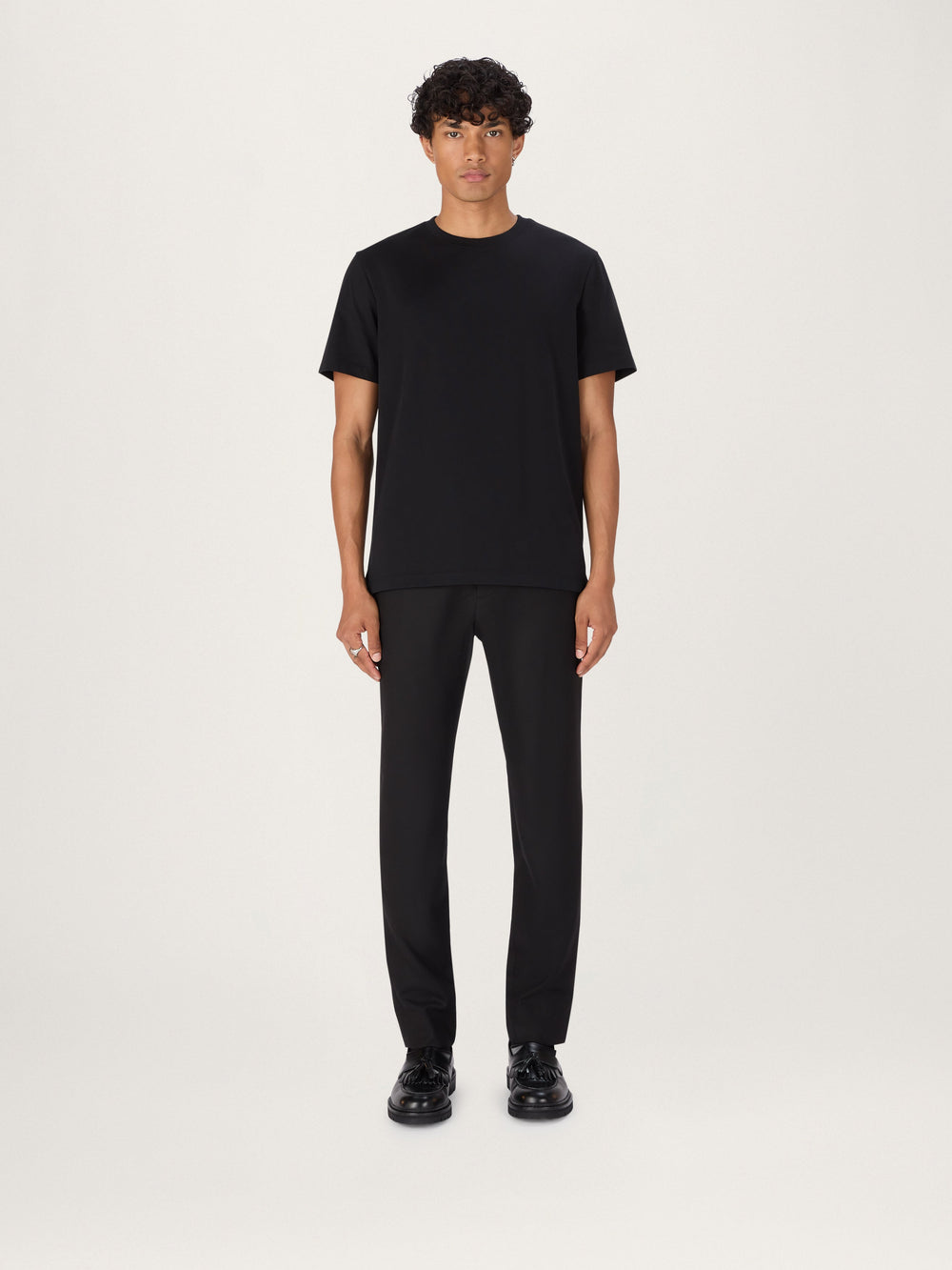 A person standing against a neutral background, wearing a black, short-sleeved, crew-neck t-shirt and dark trousers.